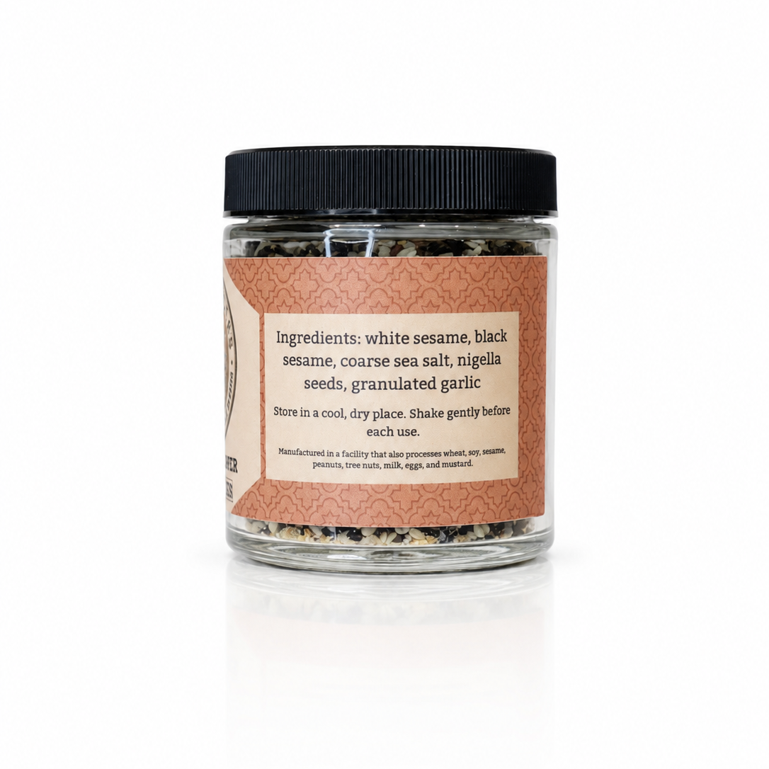 A clear glass jar of raw sea salt with a black lid and a terracotta-colored label, containing a mixture of white sesame, black sesame, coarse sea salt, nigella seeds, and granulated garlic.