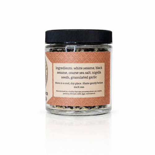 A clear glass jar of raw sea salt with a black lid and a terracotta-colored label, containing a mixture of white sesame, black sesame, coarse sea salt, nigella seeds, and granulated garlic.