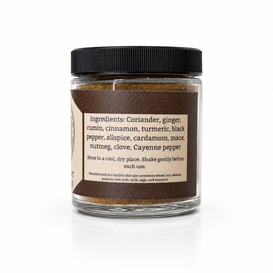 A clear glass jar of ground spice blend with a black ribbed lid and a cream label listing ingredients such as coriander, ginger, cumin, cinnamon, turmeric, black pepper, allspice, cardamom, nutmeg, clove, and cayenne pepper, shown on a white background.