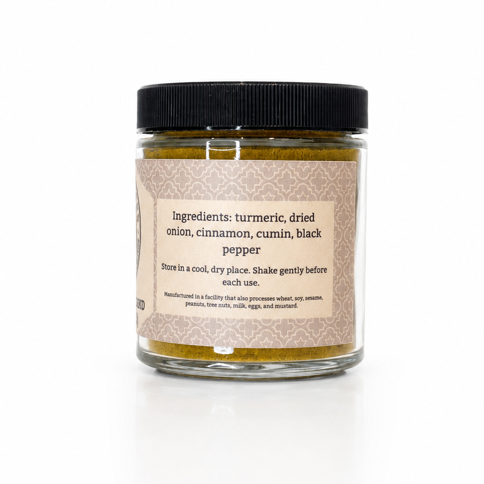 A glass jar candle with a black lid and a beige label, containing warm yellow-brown wax. The label reads 'Ingredients: turmeric, dried onion, cinnamon, cumin, black pepper' and 'Stir in a cool, dry place: Shake gently before each use.