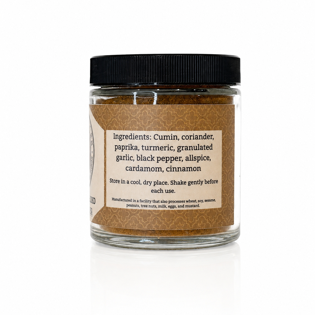A clear glass jar of ground cinnamon with a black lid and a beige label, shown on a white background with a soft reflection.